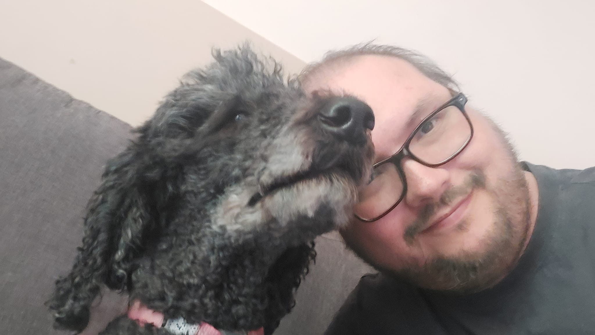 A man sits next to a black standard poodle, their heads are touching